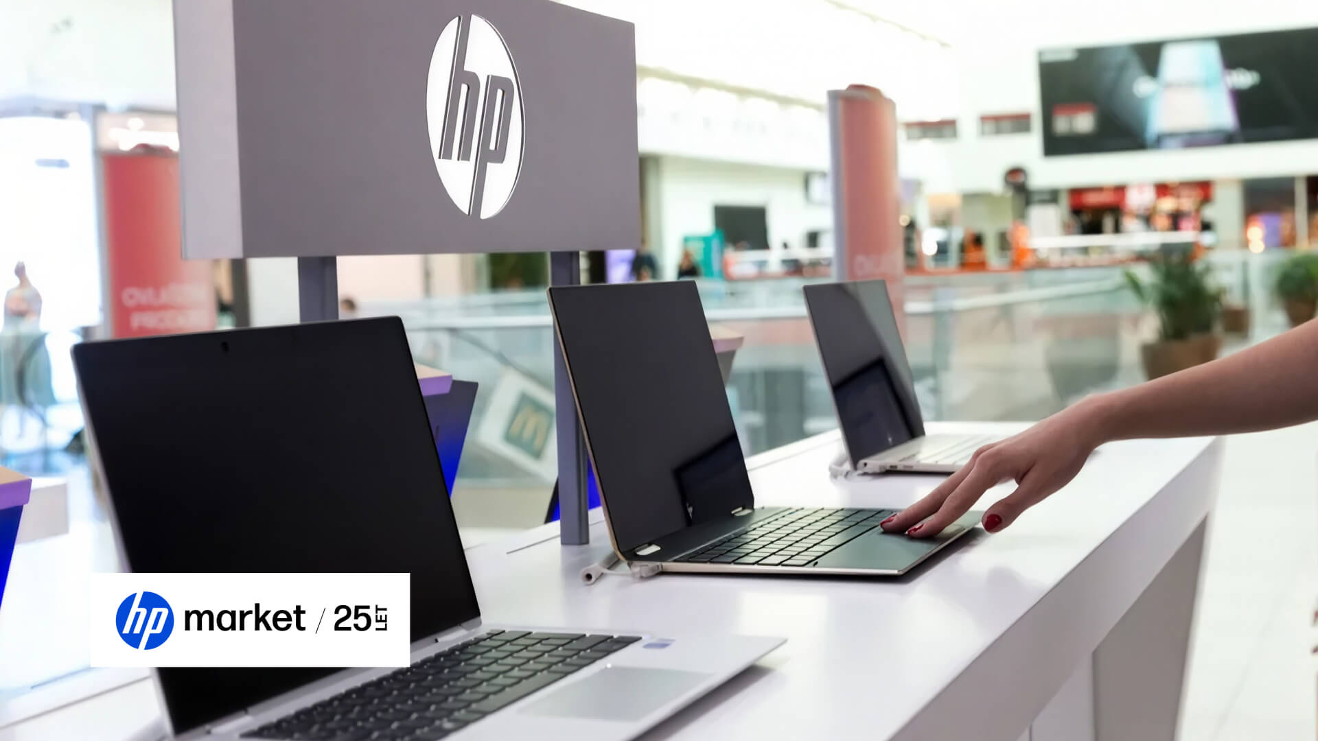 Notebooky HP vystavené na bílém pultu prodejny pod cedulí s logem HP, zákaznice zkouší jeden z nich