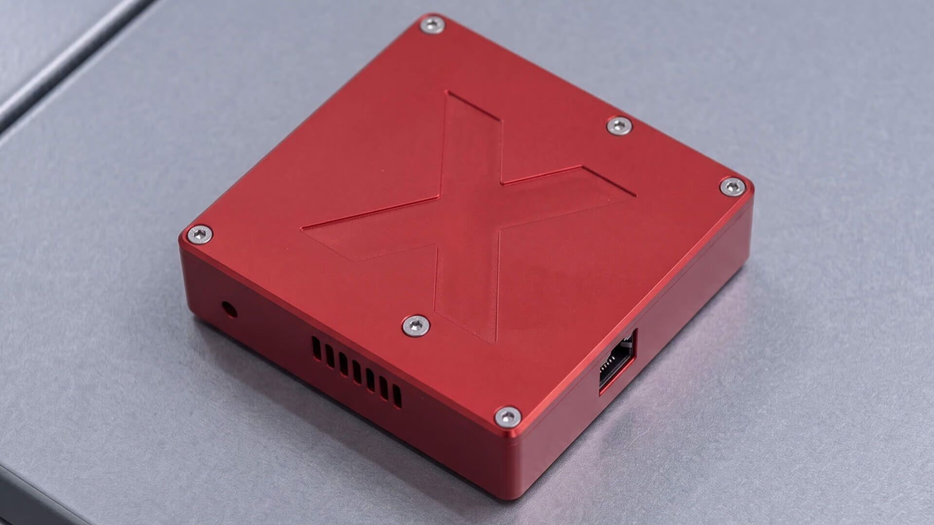 Rote SensorX-Einheit aus Aluminium mit eingeprägtem Buchstaben X, Lüftungsschlitzen und RJ45-Anschluss, auf dem grauen Blech einer Maschine liegend
